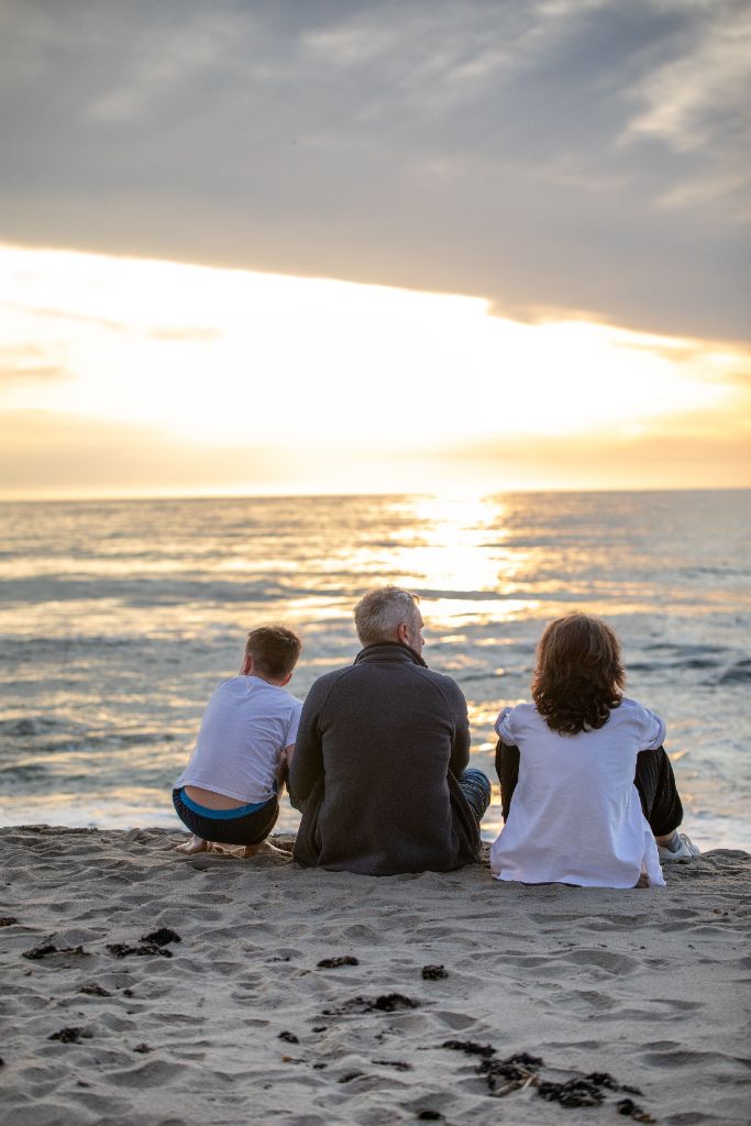 A family sits together on the sand watching the sunset — three generations at the water's edge.